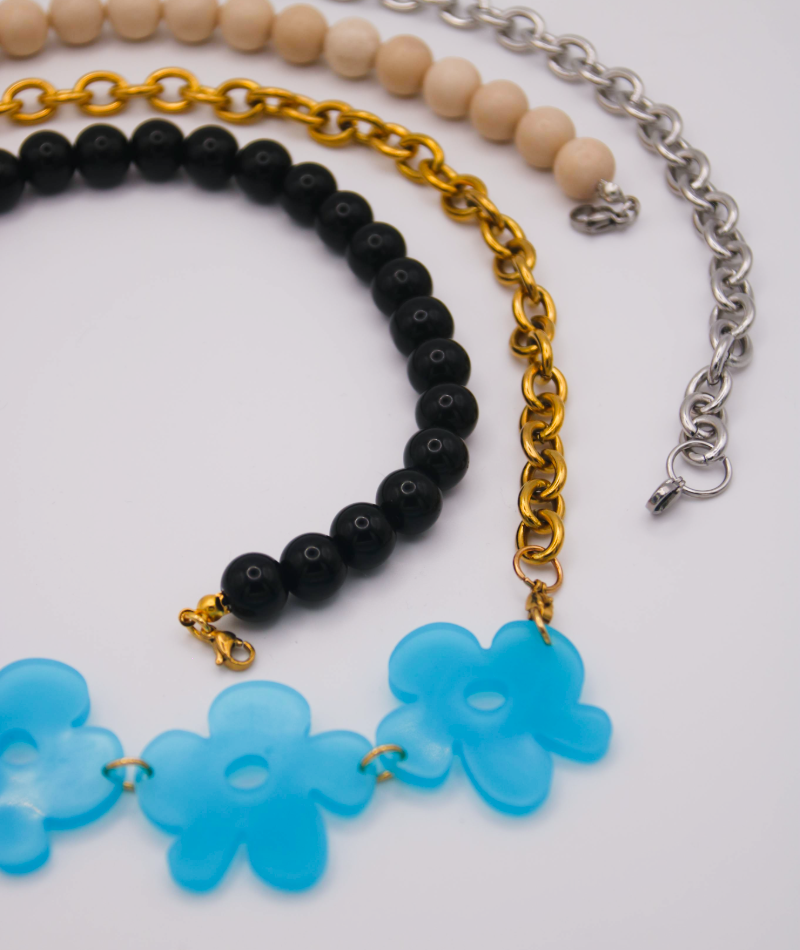 Collares intercambiables de cuentas negras y blancas, de cadenas de acero inoxidable dorado y plateado, con línea de flores en resina UV de color azul cielo. Joyería artesanal inspirada en la cultura colombiana.