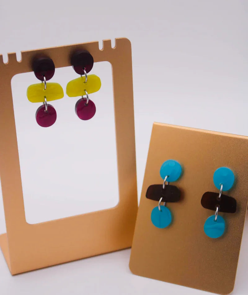 Pendientes artesanales coloridos en exhibidor dorado, joyería inspirada en cultura colombiana